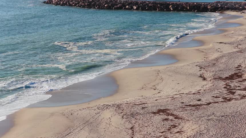 Calm ocean waves gently break in slow motion on the sandy shore next to a rock pier