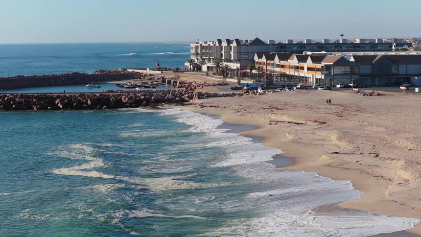 A serene aerial shot of ocean waves washing onto the sandy beach of a modern coastal town and marina