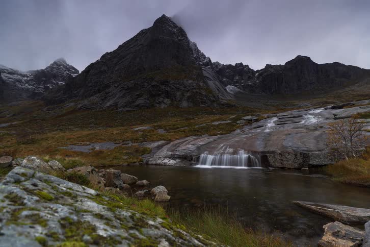 A slow-moving time-lapse reveals a waterfall cascading over smooth rocks into a tranquil pool at the foot of a towering, moody mountain