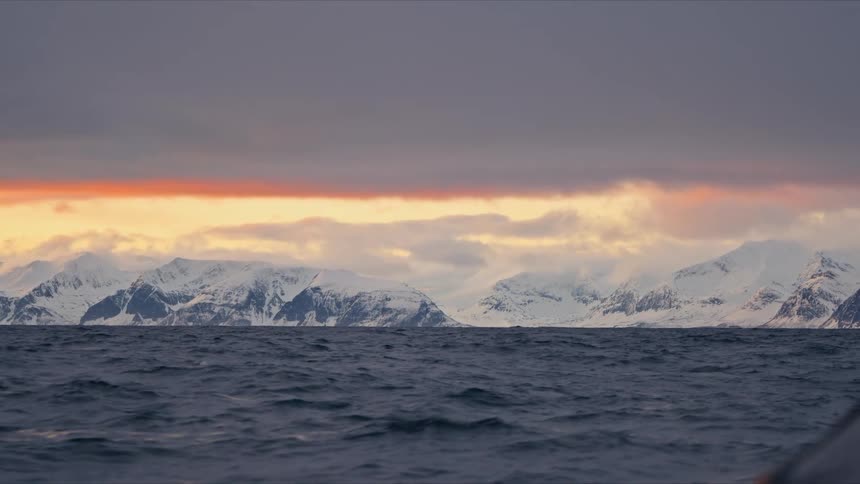 A dramatic view from a boat on a choppy Arctic sea shows snow-covered mountains against a fiery sunset on the horizon