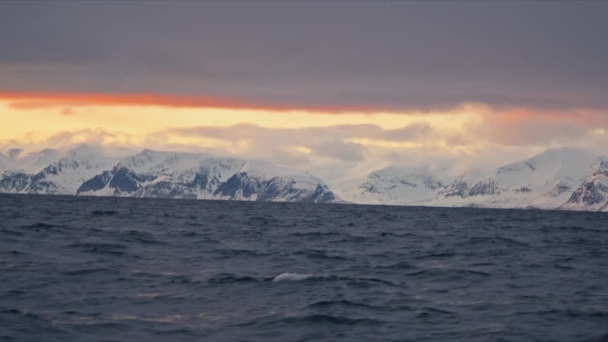 A spectacular orange sunset glows on the horizon behind a vast, snow-covered mountain range, viewed from a boat on the cold, dark sea