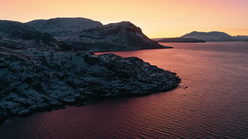 Majestic aerial view of a snowy Nordic fjord reflecting the beautiful pink and orange hues of a winter sunset