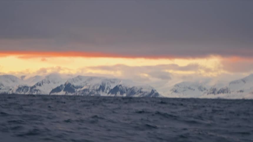 A dramatic point-of-view shot from a boat cruising through the misty, freezing Arctic ocean reveals majestic snow-covered mountains at sunset