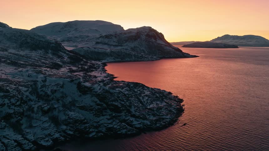 A tranquil winter sunset casts a warm, colorful glow over a snowy Norwegian fjord and its rugged coastline