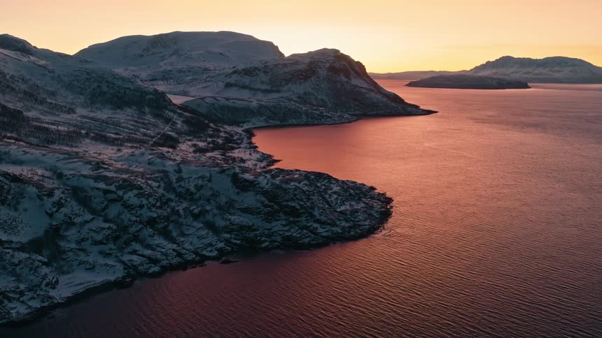 A majestic aerial view shows a tranquil winter sunset over a vast fjord, with the golden light reflecting on the water and snow-covered mountains