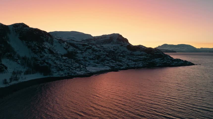 An aerial drone rises over a tranquil, snow-covered Norwegian fjord as the beautiful sunrise colors the sky and water in shades of pink and orange