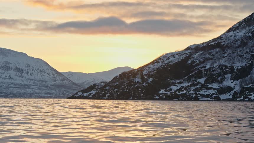 A tranquil sunset over the calm waters of a fjord with snow-covered mountains reflecting the golden light