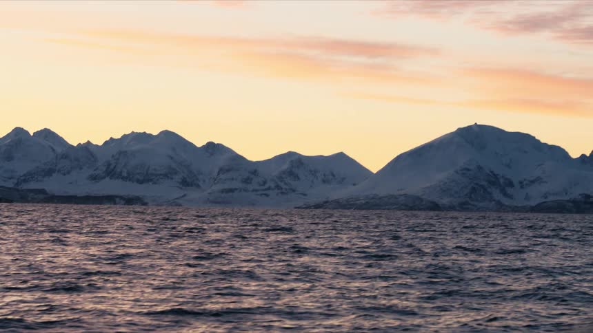 A serene view from a moving boat shows a beautiful orange and pink sunset sky over a vast, snow-covered mountain range across the rippling ocean