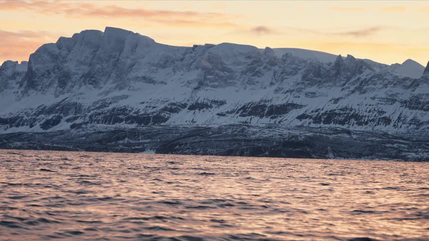 A scenic winter landscape shows snow-covered mountains along a fjord during a serene golden hour sunset