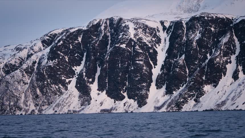 A slow zoom out showcases the immense scale of a rugged, snow-dusted mountain range bordering a tranquil fjord