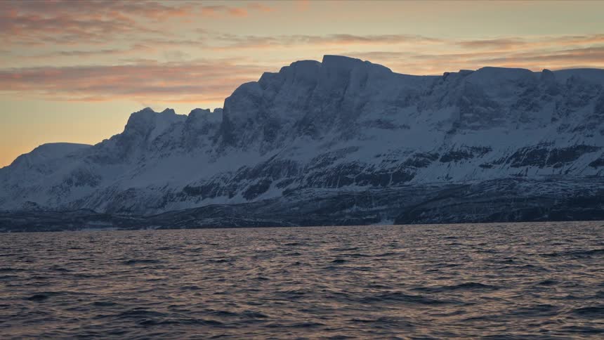 A tranquil view of majestic, snow-covered mountains across a calm fjord during a colorful sunset in winter