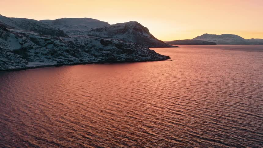 A tranquil cinemagraph captures the gentle ripples on a fjord's surface, reflecting the warm glow of a winter sunset over snow-dusted mountains