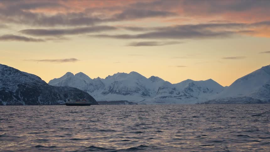 A ship sails through a tranquil arctic fjord at sunset against a stunning backdrop of snow-covered mountains