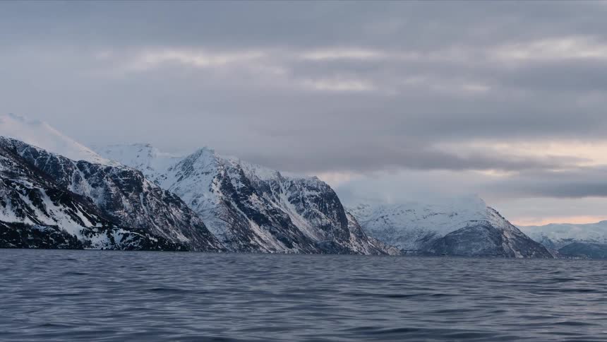 A tranquil winter scene shows gentle waves on a fjord with a majestic snow-covered mountain range under a cloudy sky