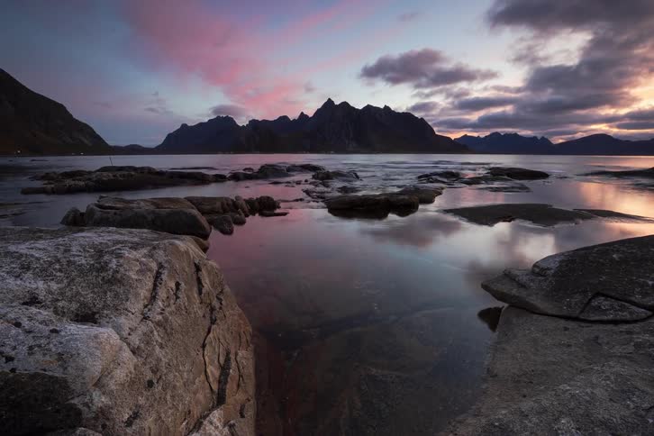 A beautiful time-lapse captures the tide coming in over a rocky shore as a vibrant pink and purple sunset reflects in the calm water
