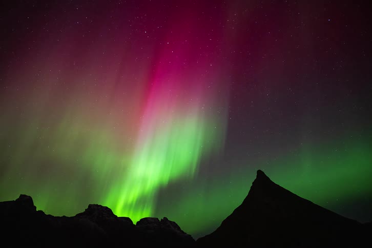 Vibrant green and magenta Northern Lights dance in a starry night sky above a silhouetted mountain peak