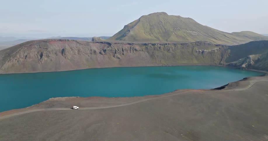 An aerial shot reveals a white SUV parked on the edge of a stunning turquoise volcanic crater lake in the remote Icelandic highlands