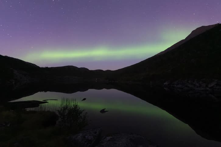 A stunning time-lapse of the green Northern Lights dancing across a purple, star-filled night sky, reflected perfectly in the still waters of a mountain lake