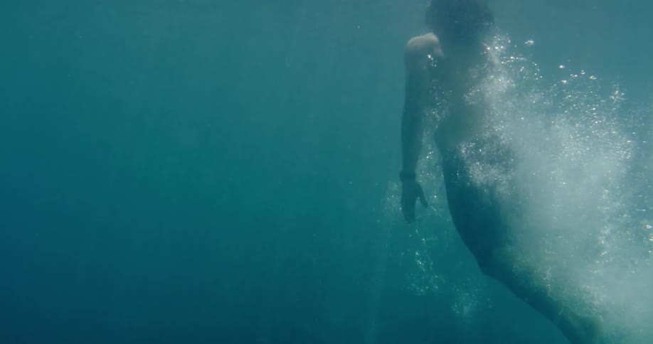 An underwater slow-motion view shows a person diving into the clear turquoise ocean, creating a trail of bubbles