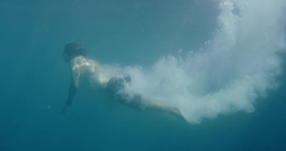 A slow-motion underwater view shows a man diving into the clear blue water, gliding forward with a trail of bubbles