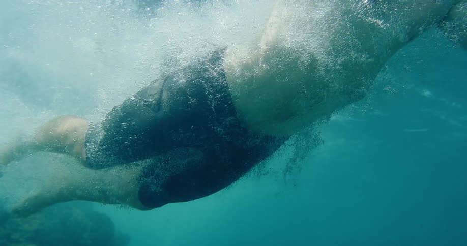 A man swims through the ocean, filmed from below the surface in slow motion