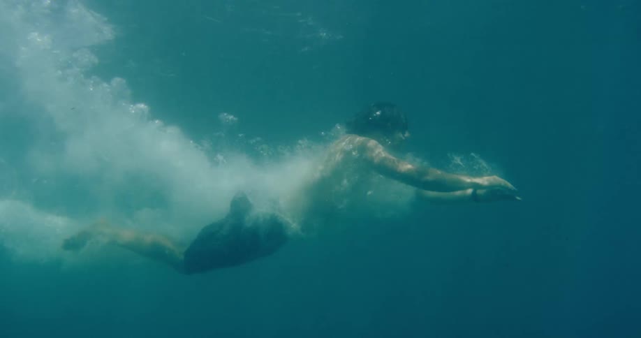 An athletic man dives into clear turquoise water, gliding gracefully underwater in slow motion surrounded by a trail of bubbles