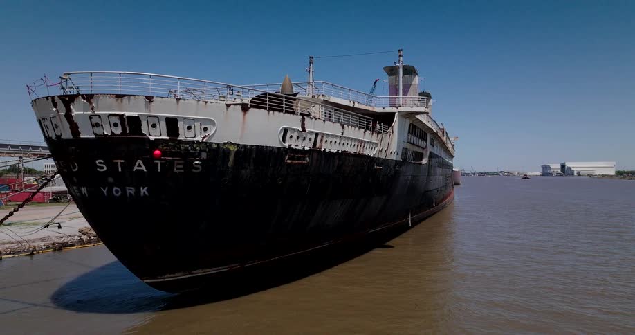 An aerial view starts at the bow of the historic and rusty SS United States and slowly pulls back to reveal the massive ocean liner docked in an industrial harbor