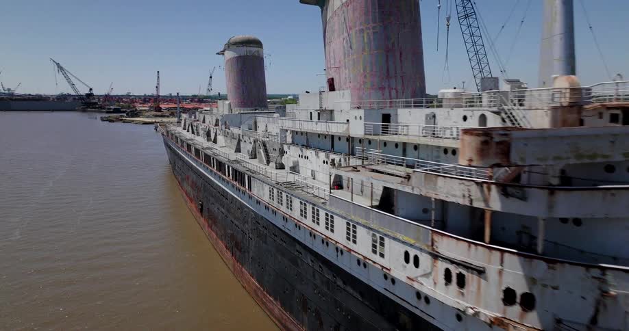 An aerial fly-by reveals the massive, decaying hull of the historic SS United States ocean liner, rusting away while docked in a busy industrial harbor