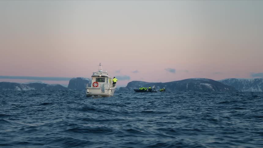A small tour boat and a zodiac float on the choppy arctic ocean at dusk with snowy mountains on the horizon