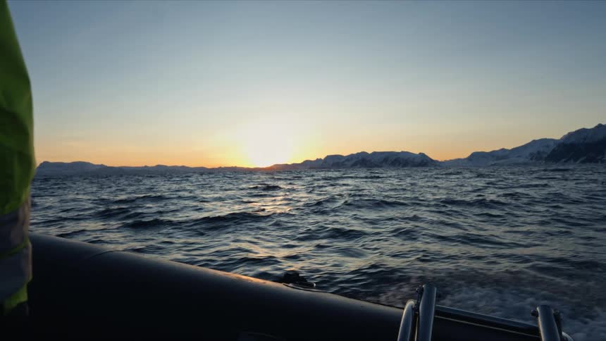 A RIB boat speeds across the choppy arctic ocean at sunset, with water spraying against the backdrop of distant snow-covered mountains