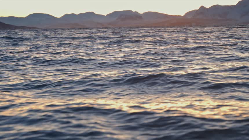 Gentle ocean waves ripple in slow motion at sunset with a snow-covered mountain range in the distance