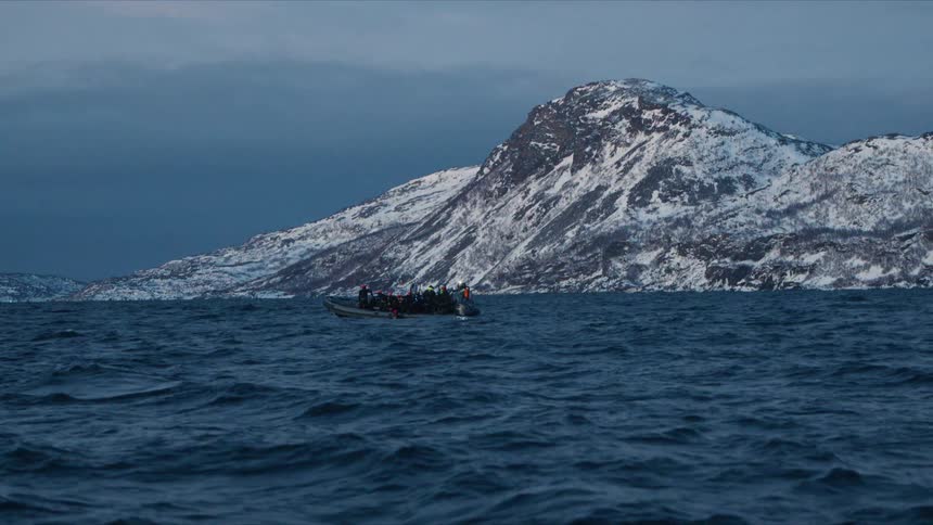 Tourists on a rigid inflatable boat float on choppy arctic waters with a dramatic snow-covered mountain in the background
