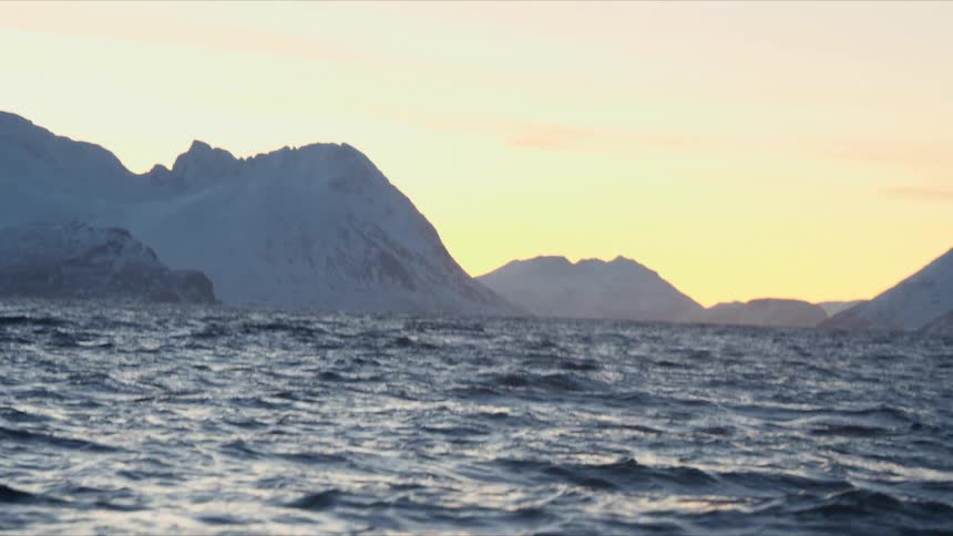 Calm ocean waves roll through a majestic fjord surrounded by snow-capped mountains at sunset
