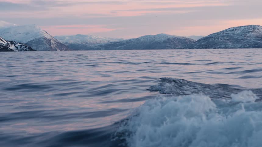 A slow motion boat ride through a tranquil Norwegian fjord reveals a stunning winter landscape of snow-capped mountains at sunset