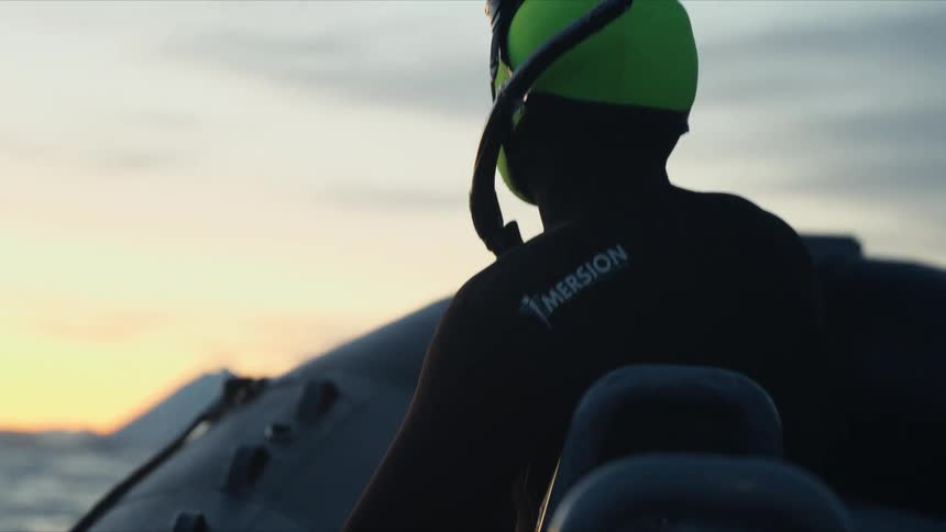 A person in a wetsuit on a boat watches a whale surface in the arctic waters at sunset in slow motion