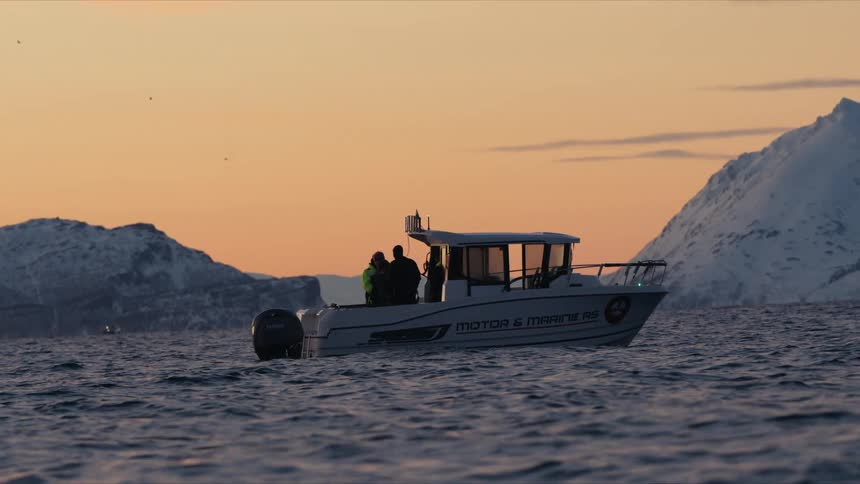 A motorboat with silhouetted passengers drifts on the water at sunset, with snowy mountains creating a stunning backdrop
