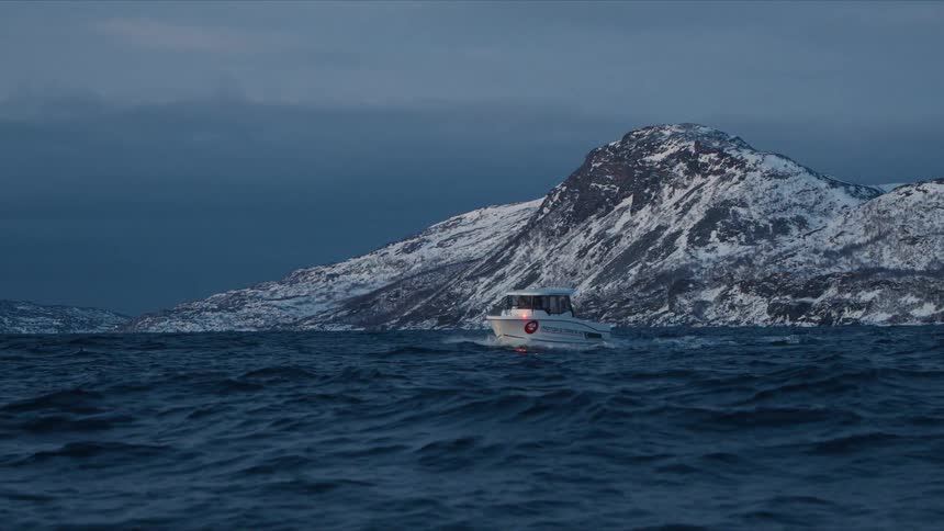 A motorboat sails through the dark, choppy sea in slow motion at dusk, with snow-covered mountains in the background