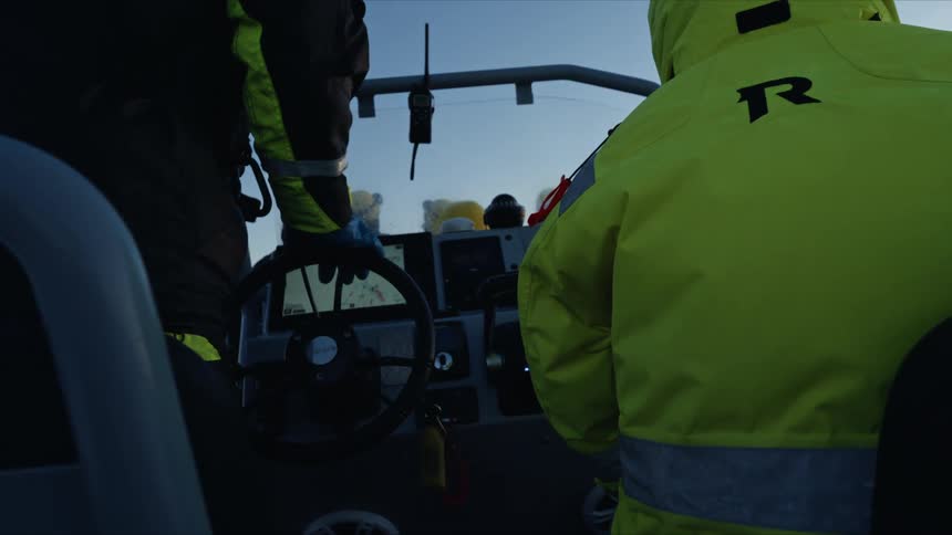 A boat captain and crew member navigate the open sea at dusk, with the focus on the helm and electronic navigation screen