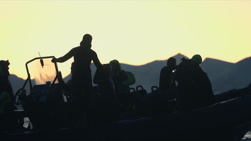 An expedition team in a rigid inflatable boat is silhouetted against a golden sunrise in a cold, mountainous arctic environment