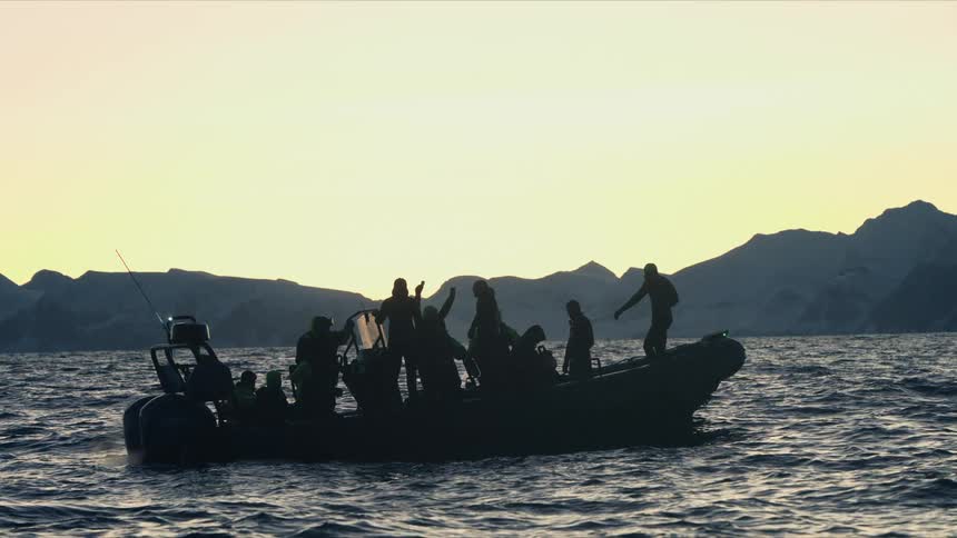 A silhouette of a group of tourists on a rigid inflatable boat floats on the calm arctic sea at sunset, with distant snowy mountains on the horizon