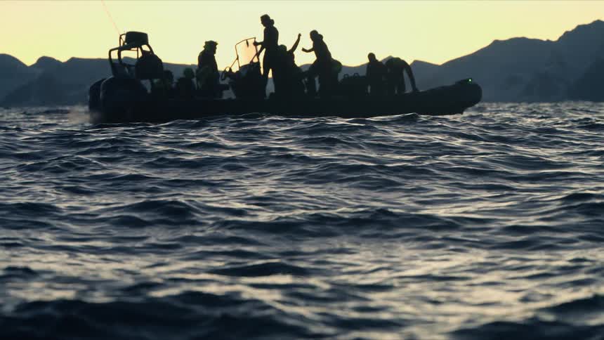 A silhouetted expedition boat with explorers floats on the open water at sunset before the focus shifts to the waves in the foreground