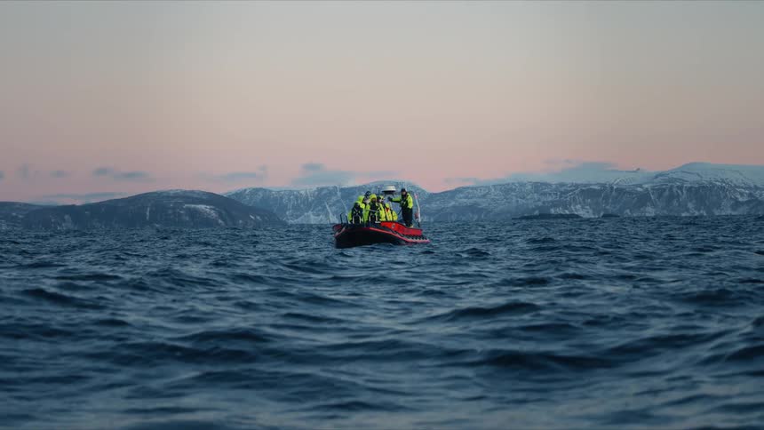 A rigid inflatable boat with a tour group bobs on the choppy arctic ocean at dusk with a beautiful snowy mountain range behind them