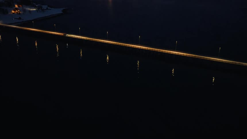 Rising aerial shot follows a car driving over a long, illuminated bridge at night towards a snowy, coastal town