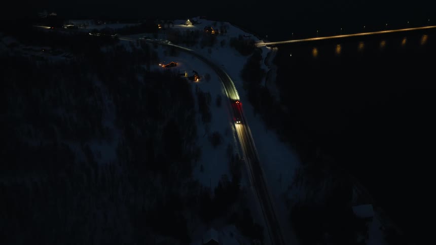 A serene aerial perspective shows cars navigating a winding, snow-covered road along the water's edge at night