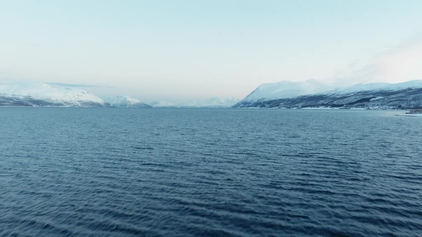 A sweeping aerial shot glides over the choppy waters of a northern fjord, tilting up to reveal a vast, serene landscape of snow-covered mountains