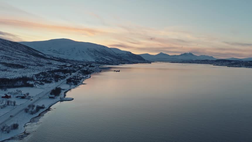 An aerial view of a serene Nordic fjord at sunset, with a snowy coastline, mountains, and a quiet road with passing cars