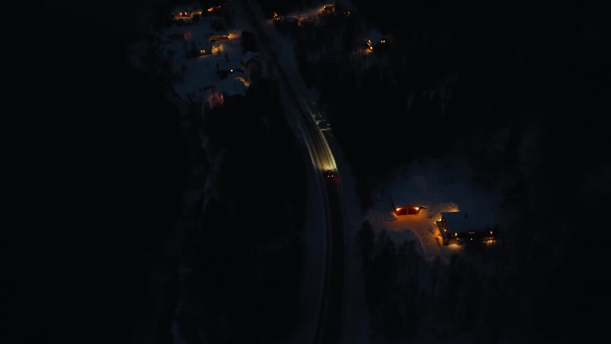 Aerial top-down view of cars driving on a dark, snowy road at night, pulling back to reveal a coastal village and mountains at dusk