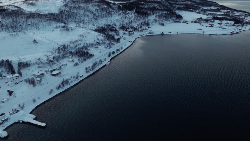 A serene aerial drone shot flies over a dark fjord, revealing a snow-covered village nestled along the wintry shoreline