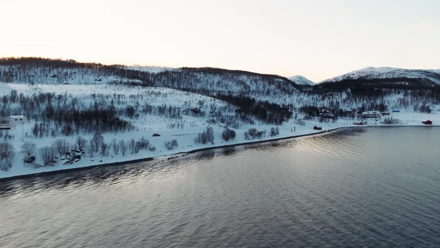 An aerial view of a tranquil, snow-covered village on the edge of a fjord during a Nordic winter sunset