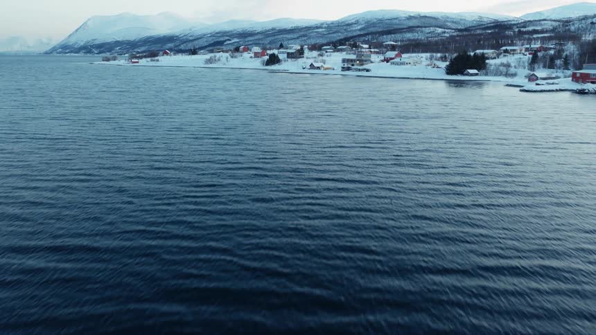 An aerial shot flies backwards over the calm, dark blue water, revealing a snowy village on the shore of a Norwegian fjord with mountains in the background